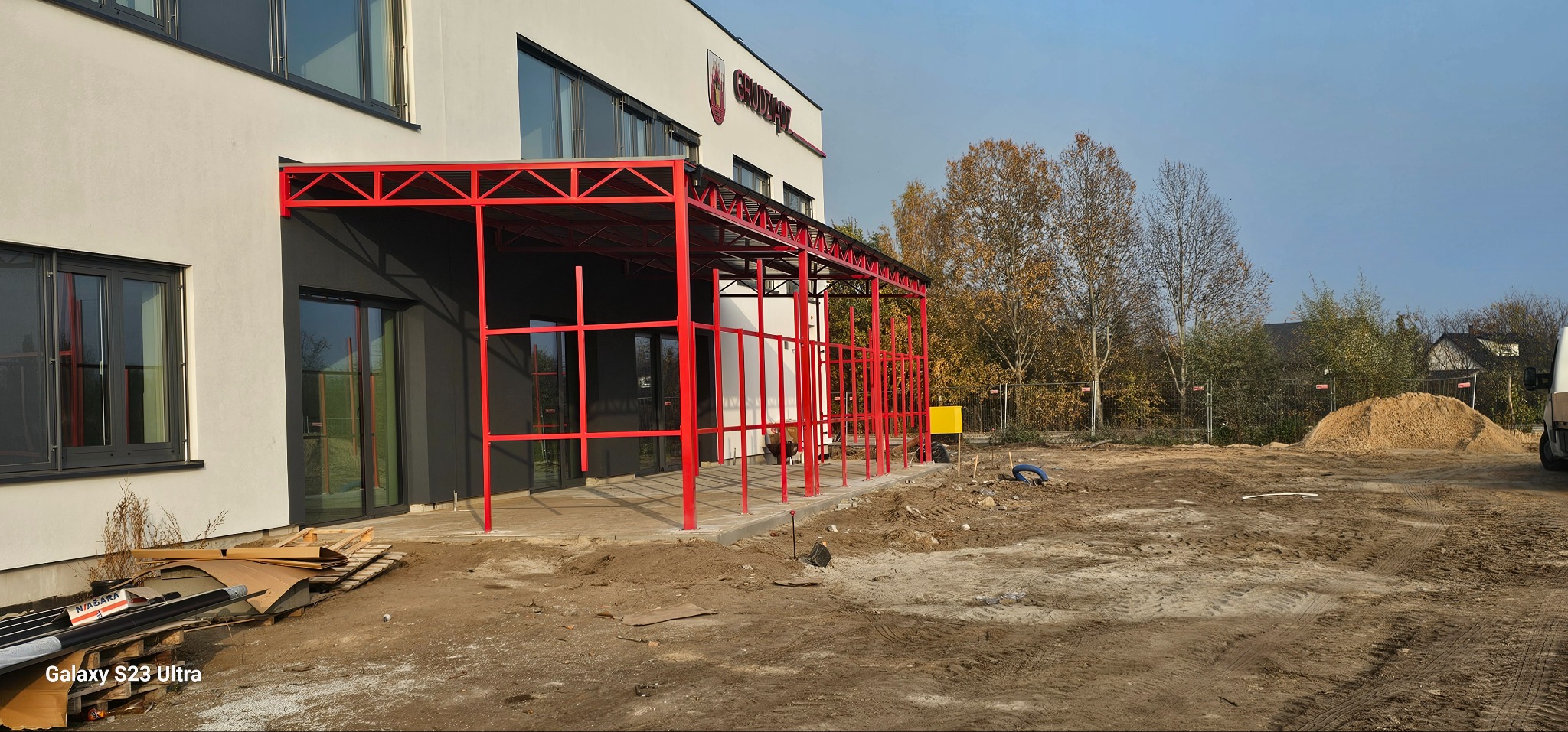 Czerwona konstrukcja stalowa zadaszenia przy budynku z oknami. Widoczne materiały budowlane i teren budowy. Elementy spawane, nowoczesny design.