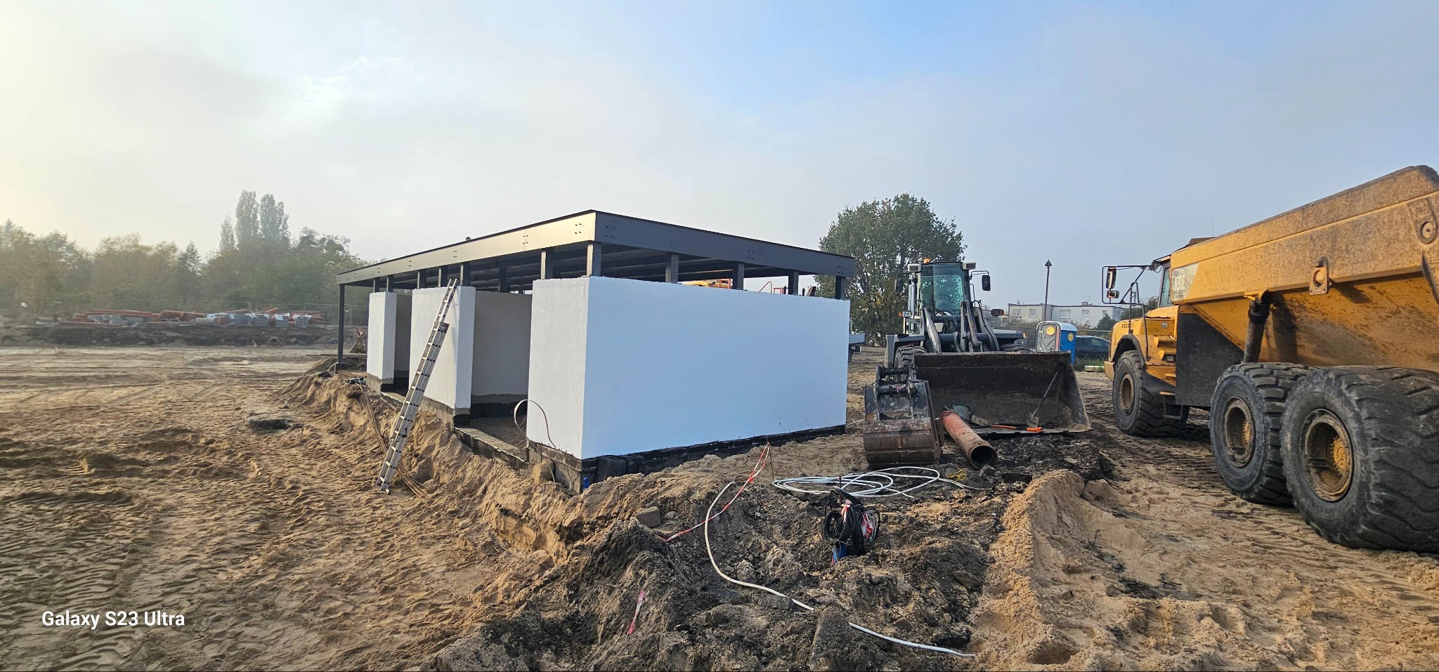 Wczesny etap budowy hali z metalową konstrukcją i białymi ścianami. Na pierwszym planie ziemia, w tle maszyny budowlane i drzewa. Widoczne kable i drabina.