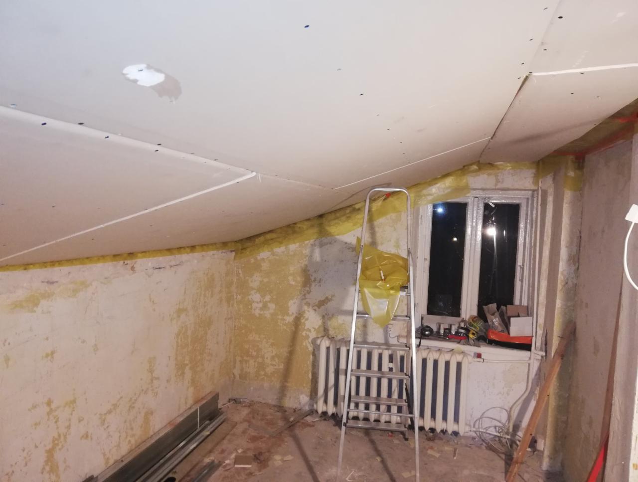 Wnętrze remontowanego poddasza z oknem: częściowo zamontowane płyty gipsowe na skośnym suficie, drabina, grzejnik pod oknem, ściany z resztkami starej farby i żółtą izolacją.