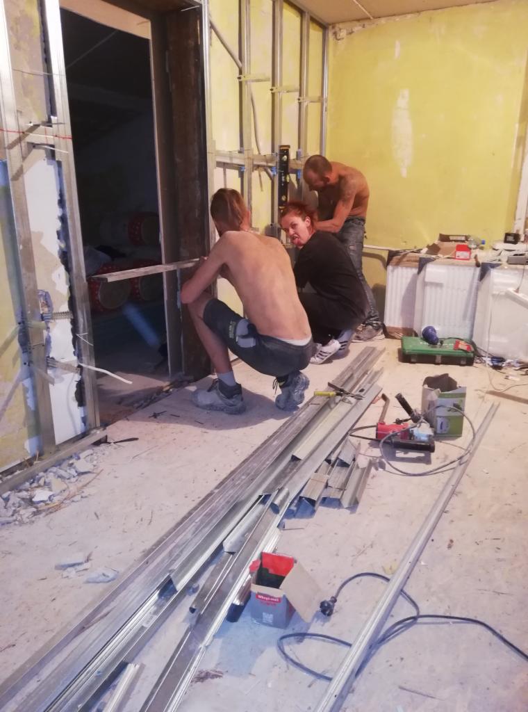 Trzech mężczyzn w trakcie rozbudowy domu: montaż metalowych profili, widoczne narzędzia i materiały budowlane, surowy stan wykończenia wnętrza.