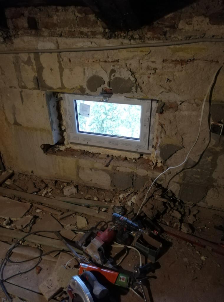 Montaż okna PCV w surowym murze, widoczne narzędzia budowlane na podłodze, pianka montażowa wokół ramy okiennej.