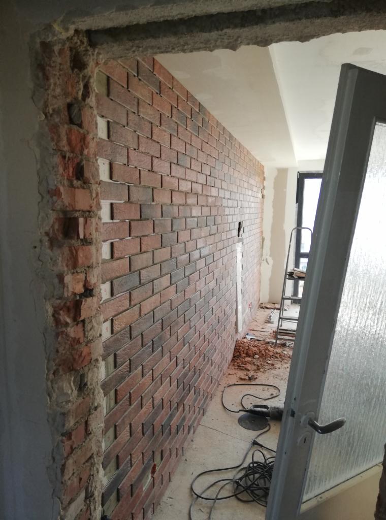 Wnętrze w trakcie prac: ściana wyłożona cegłą klinkierową w odcieniach czerwieni i brązu, widoczny fragment surowej ściany z widoczną strukturą cegieł przy otworze drzwiowym, na podłodze gruz...