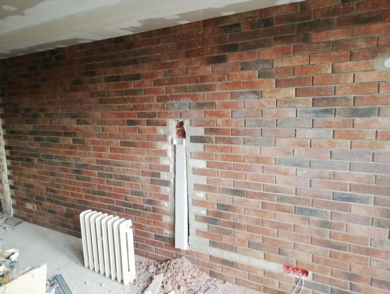 Ściana z cegły klinkierowej w trakcie montażu, widoczne niedokończone fragmenty, grzejnik i instalacja elektryczna.