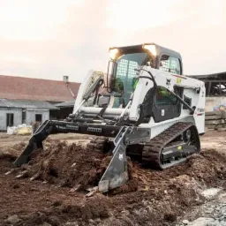 Biała ładowarka gąsienicowa Bobcat z osprzętem do spulchniania ziemi pracuje na placu budowy, w tle budynki gospodarcze z ceglanym dachem.