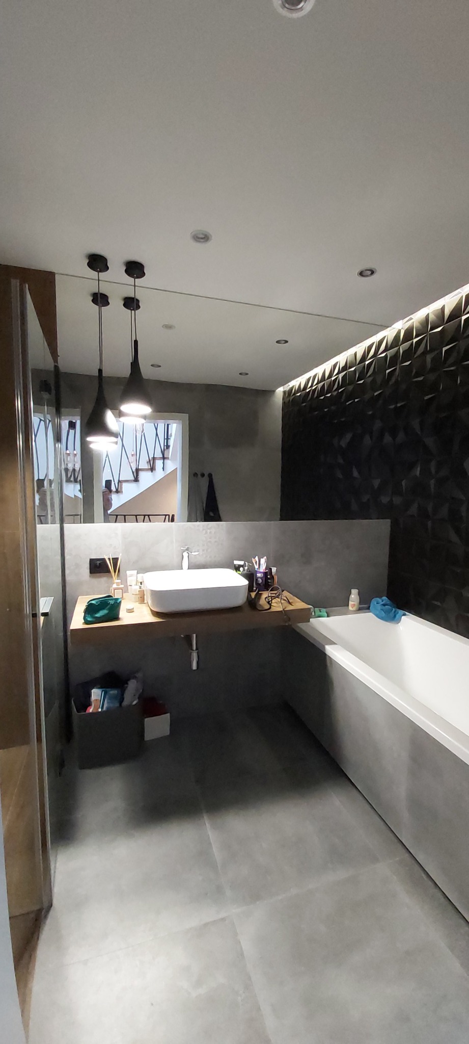 Nowoczesna łazienka z umywalką nablatową na drewnianym blacie, geometryczną ścianą z podświetleniem LED i widoczną w lustrze klatką schodową.