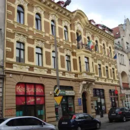 Ulica z kamienicami w stylu secesyjnym, z flagami Włoch, Unii Europejskiej i Polski powiewającymi na balkonie jednej z nich. Na parterze widoczna księgarnia i lokal gastronomiczny.