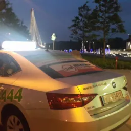 Biała taksówka Skoda z numerem 6444444 zaparkowana nocą przy fontannie w mieście, widoczne naklejki reklamowe na tylnej szybie i lampa taksówkarska na dachu.