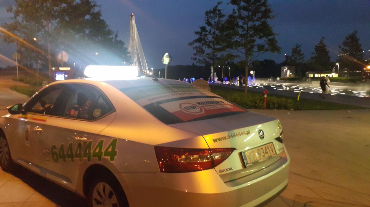 Biała taksówka Skoda z numerem 6444444 zaparkowana nocą przy fontannie w mieście, widoczne naklejki reklamowe na tylnej szybie i lampa taksówkarska na dachu.