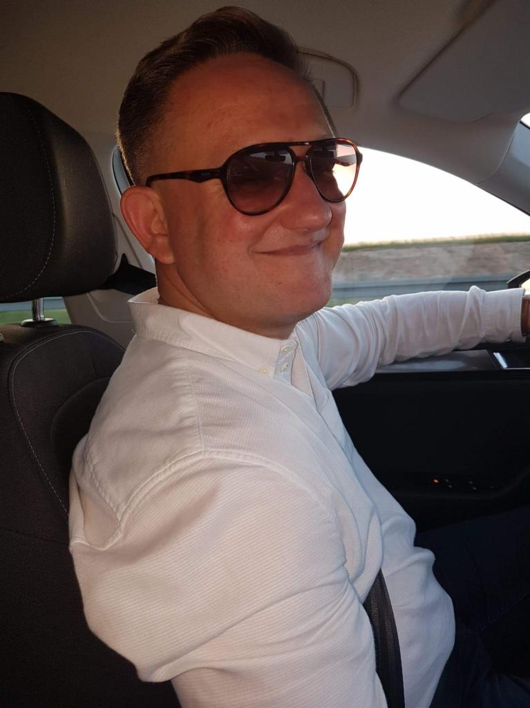 Uśmiechnięty mężczyzna w okularach za kierownicą samochodu, w trakcie jazdy. Widoczny pas bezpieczeństwa i fragment deski rozdzielczej.