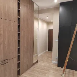 Nowoczesna garderoba z drewnopodobnymi frontami i lustrem, widoczna drabina oparta o czarną szafę w przedpokoju z oświetleniem punktowym.