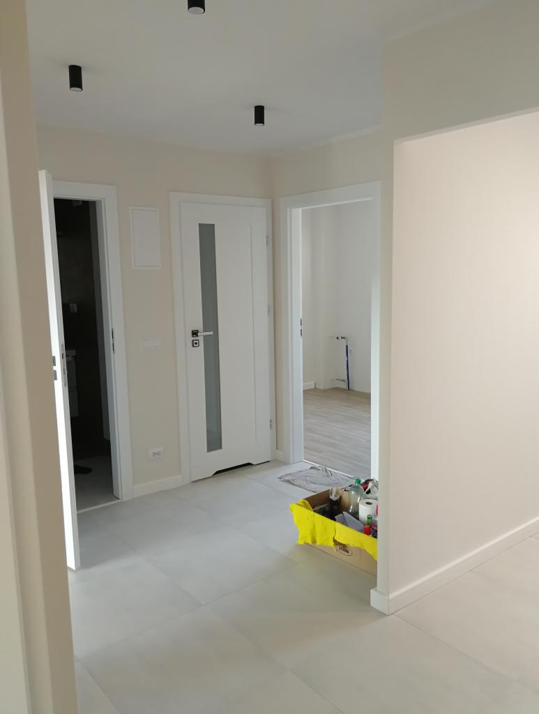 Wnętrze mieszkania w trakcie odświeżania: świeżo położone płytki podłogowe, białe drzwi z przeszkleniem, karton z materiałami budowlanymi w rogu, widok na kolejne pomieszczenia.