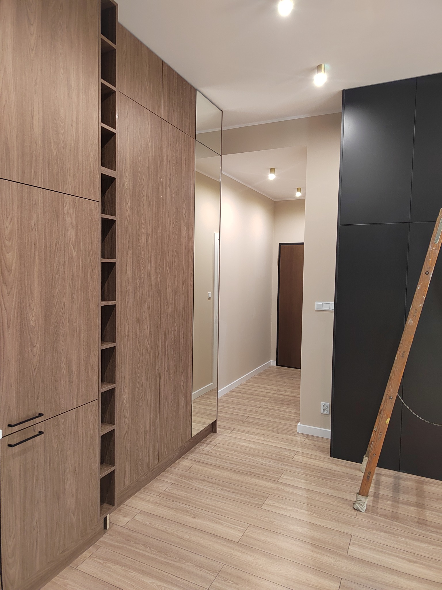 Nowoczesna garderoba z drewnopodobnymi frontami i lustrem, widoczna drabina oparta o czarną szafę w przedpokoju z oświetleniem punktowym.