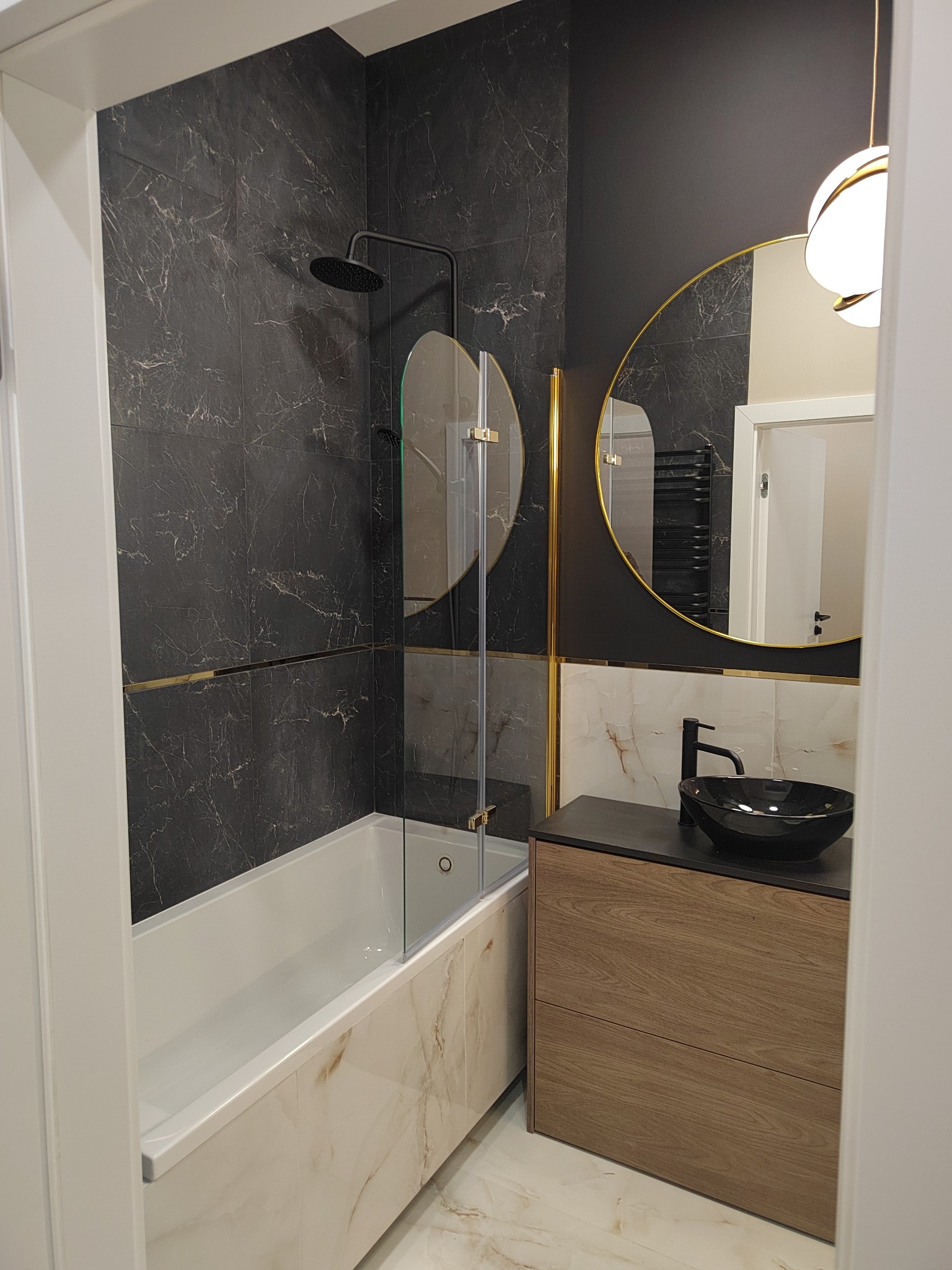 Nowoczesna łazienka z czarnymi marmurowymi płytkami, kabiną prysznicową nad wanną, okrągłym lustrem w złotej ramie i czarną umywalką nablatową.