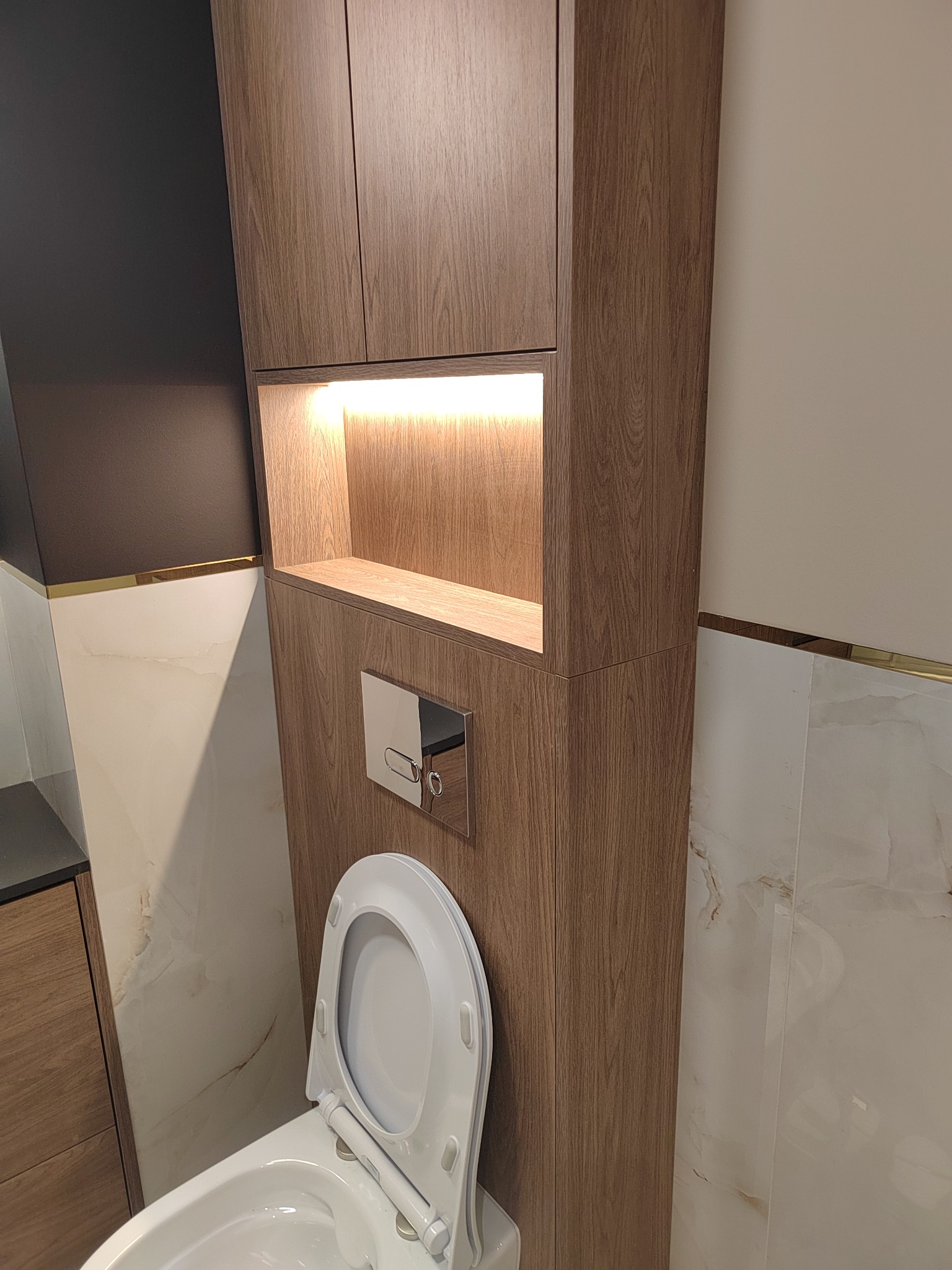 Nowoczesna toaleta wisząca z zabudową z drewnopodobnych szafek z oświetleniem LED i chromowanym przyciskiem spłukiwania, wykończona marmurowymi płytkami z delikatną złotą listwą.