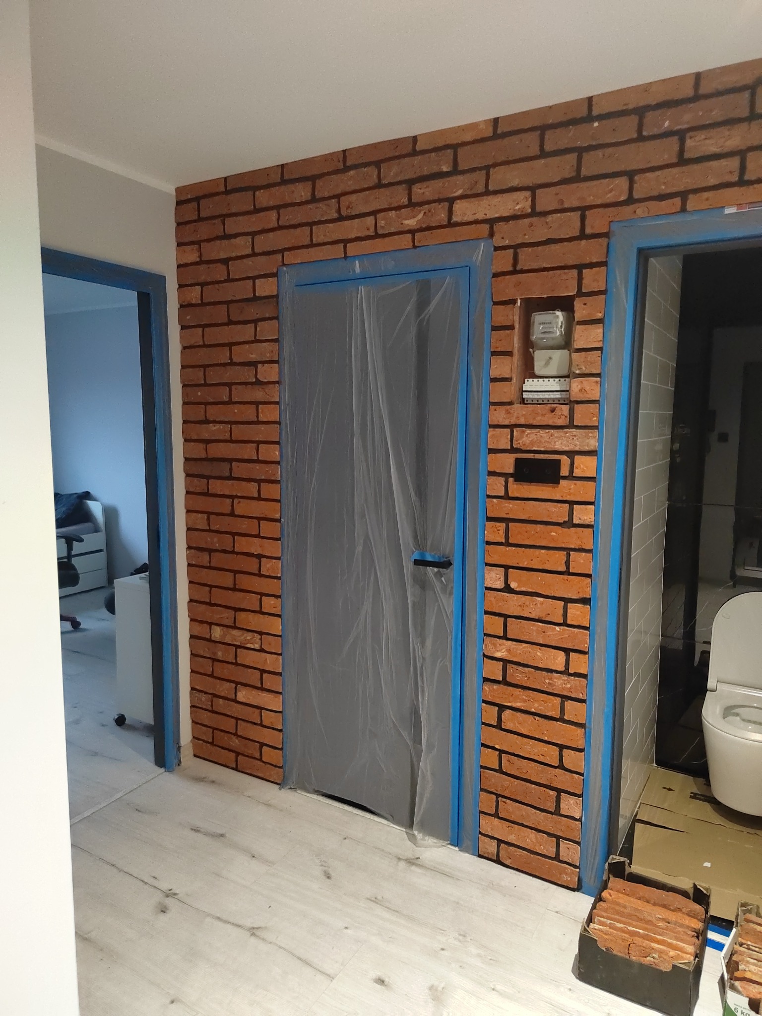 Ściana wykończona cegłą klinkierową, z zabezpieczonymi folią i taśmą malarską drzwiami, widoczny licznik elektryczny i fragment łazienki z toaletą. Na podłodze karton z cegłami.