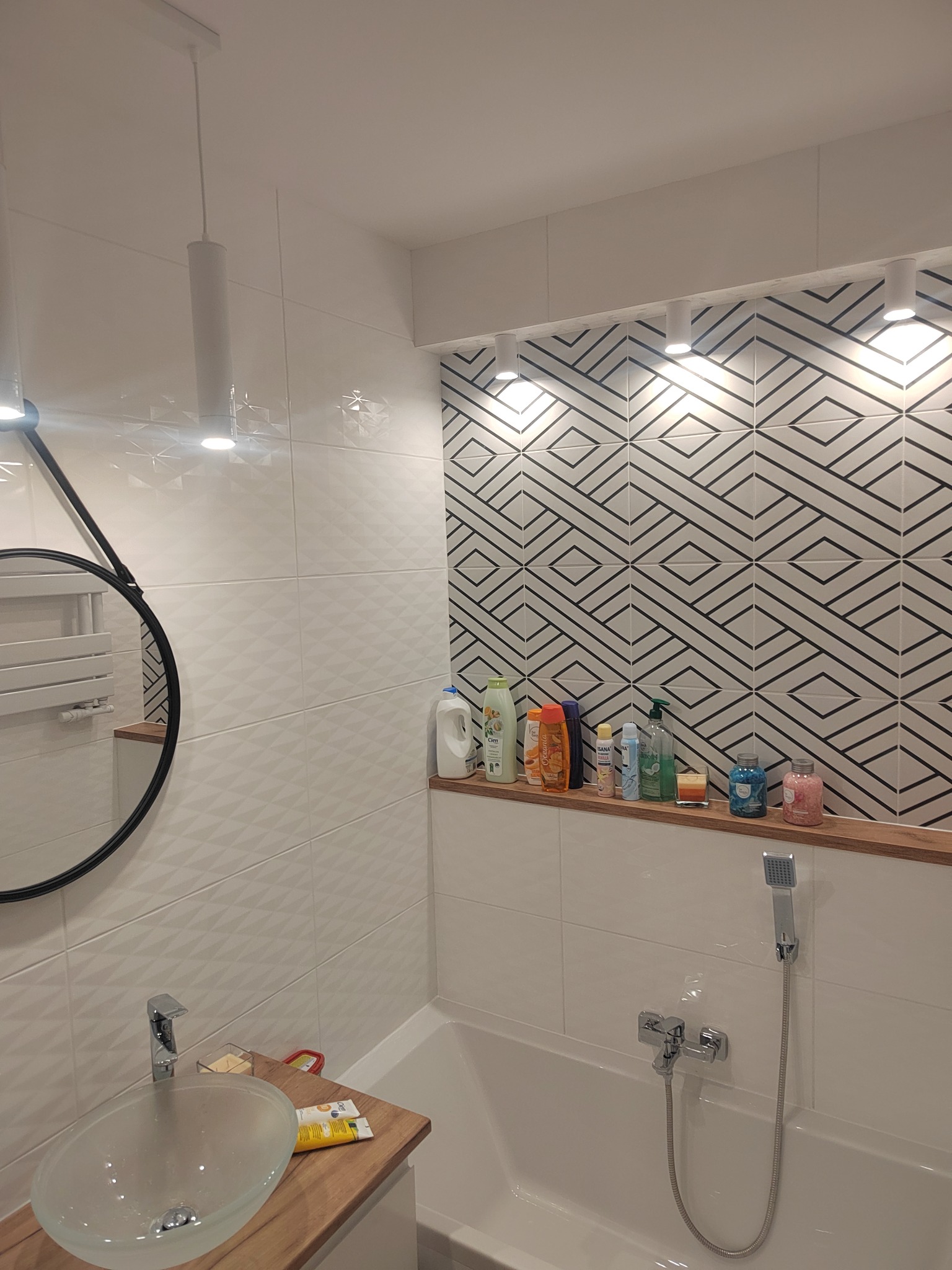 Nowoczesna łazienka z umywalką na drewnianym blacie, geometrycznymi płytkami nad wanną i podświetlaną półką z kosmetykami.