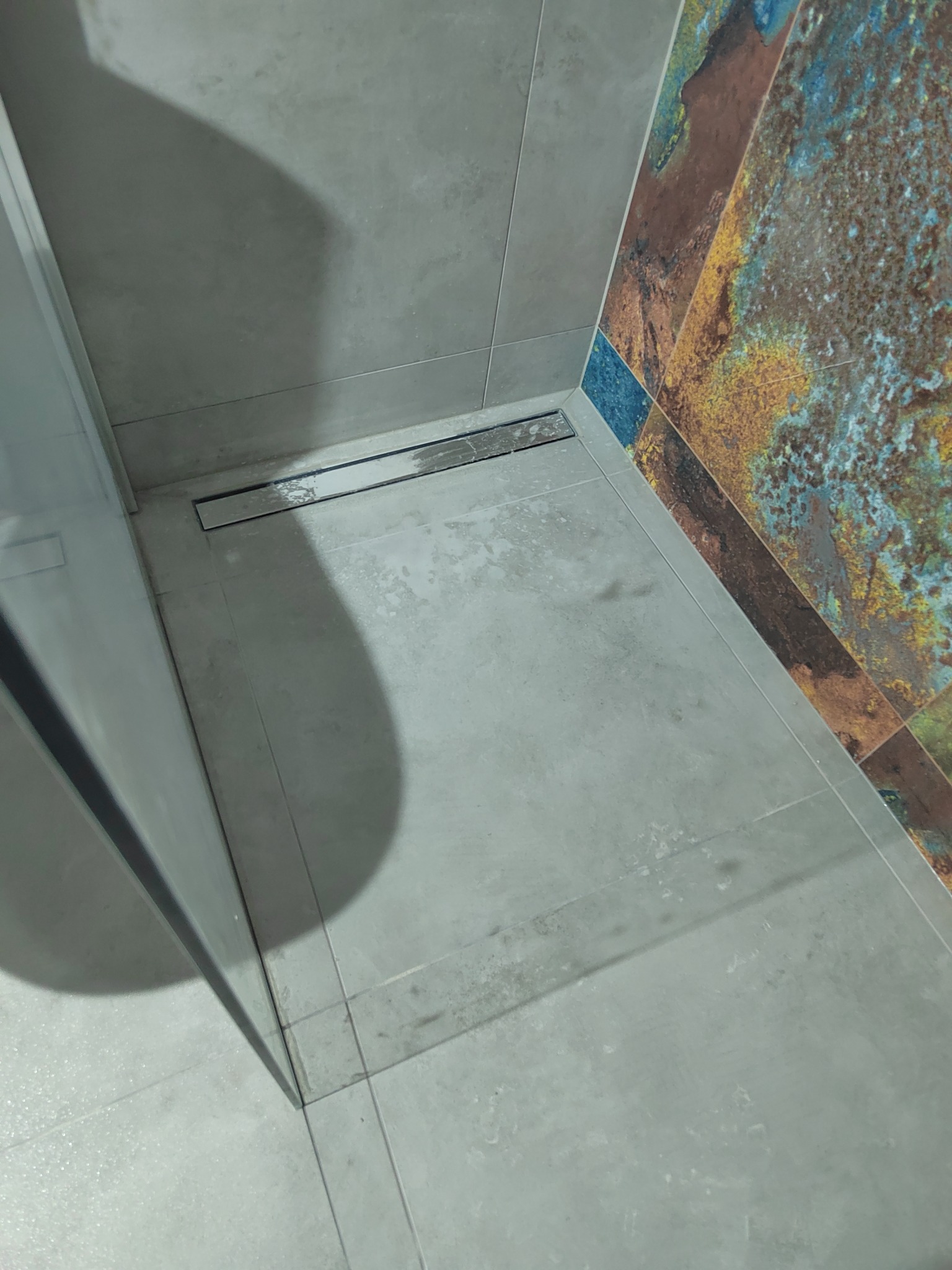 Nowoczesny odpływ liniowy w kabinie prysznicowej z szarymi płytkami podłogowymi i ścianą wyłożoną płytkami w kolorach ziemi i błękitu, widok z góry.