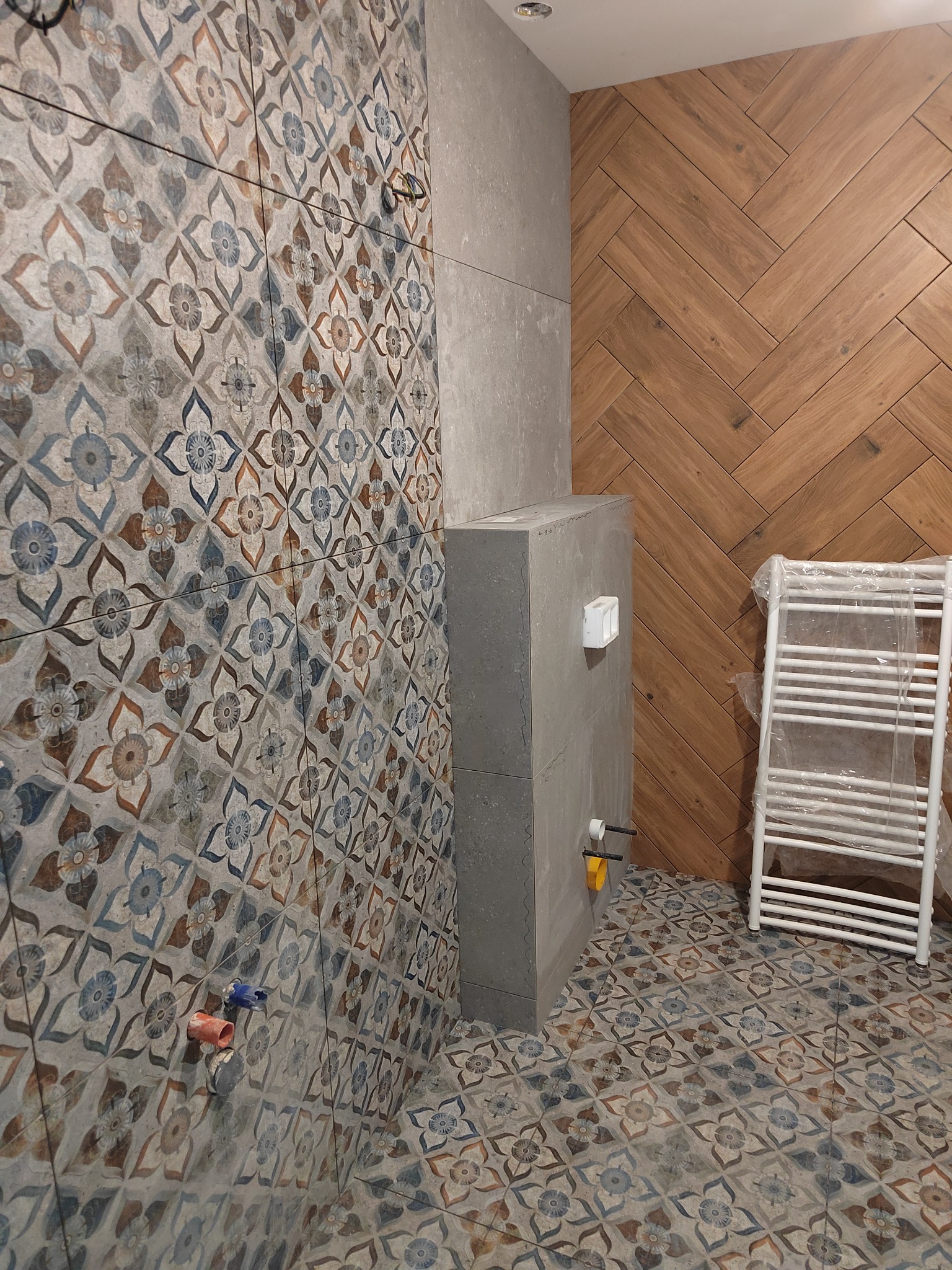 Łazienka w trakcie remontu: ściana wyłożona wzorzystymi płytkami ceramicznymi, drewnopodobna ściana ułożona w jodełkę, zabudowa stelaża WC z szarej płyty, grzejnik drabinkowy zabezpieczony folią.