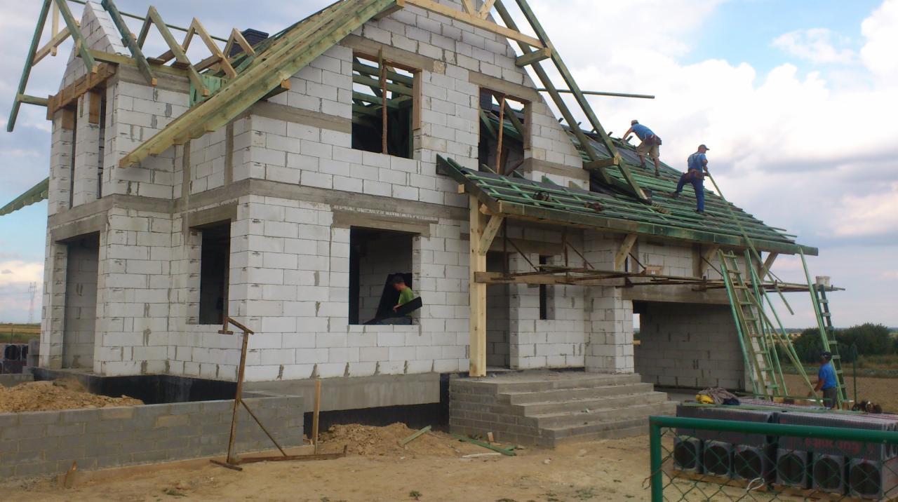 Dwukondygnacyjny dom murowany w trakcie budowy z białych bloczków, widoczni robotnicy na dachu układający zieloną membranę dachową, szare niebo w tle.