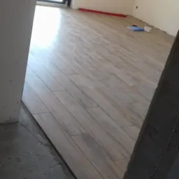 Ułożona podłoga z płytek imitujących drewno w jasnym pomieszczeniu, widoczny poziomica i opakowanie po płytkach, ściany przygotowane do malowania.