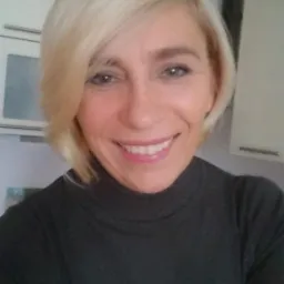 Uśmiechnięta blond kobieta w ciemnym golfie, selfie w kuchni z szafkami w tle i czajnikiem na górnej półce. Naturalny makijaż, krótka fryzura.