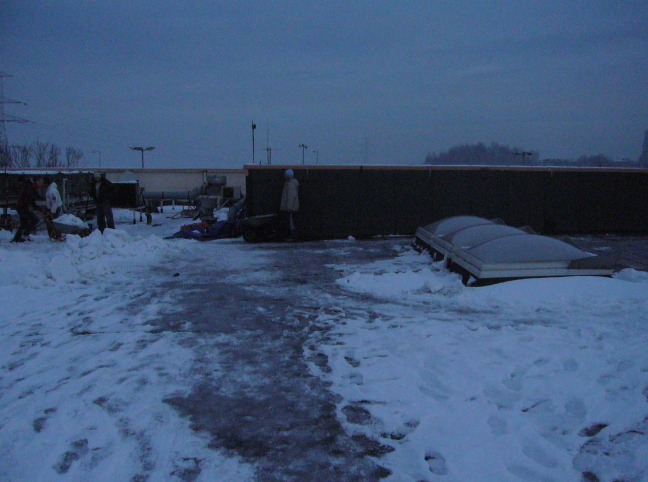 Ekipa usuwa śnieg z dachu budynku w pochmurny dzień, widoczne odśnieżone fragmenty oraz narzędzia pracy.