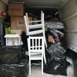 Wnętrze furgonetki załadowane kartonami, meblami i workami, w tym dwa białe krzesła na pierwszym planie, roślina doniczkowa w kartonie i pudełka marki Kemon na górze.