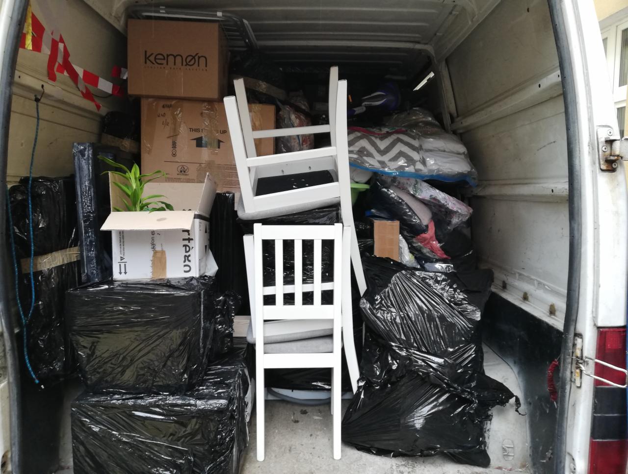 Wnętrze furgonetki załadowane kartonami, meblami i workami, w tym dwa białe krzesła na pierwszym planie, roślina doniczkowa w kartonie i pudełka marki Kemon na górze.
