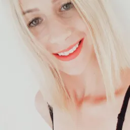 Uśmiechnięta blondynka z intensywnie czerwoną szminką, w czarnej bieliźnie na tle białej ściany. Ujęcie z dołu, eksponujące twarz i dekolt.