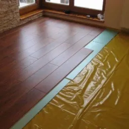 Układanie paneli podłogowych z brązowego laminatu, widoczna warstwa izolacyjna i folia ochronna na podłodze, fragment ściany z cegły dekoracyjnej.