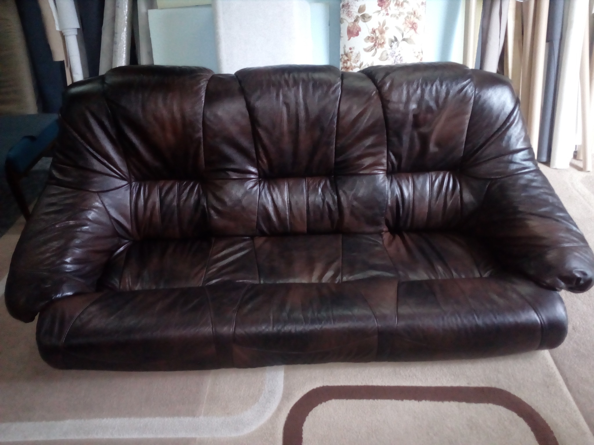 Ciemnobrązowa skórzana sofa trzyosobowa z widocznymi śladami użytkowania, ustawiona na dywanie z geometrycznym wzorem.