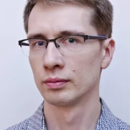 Mężczyzna w okularach z brązowymi włosami, ubrany w koszulę w kratę, na jasnym tle. Portret z bliska, skupiający się na twarzy i wyrazie.