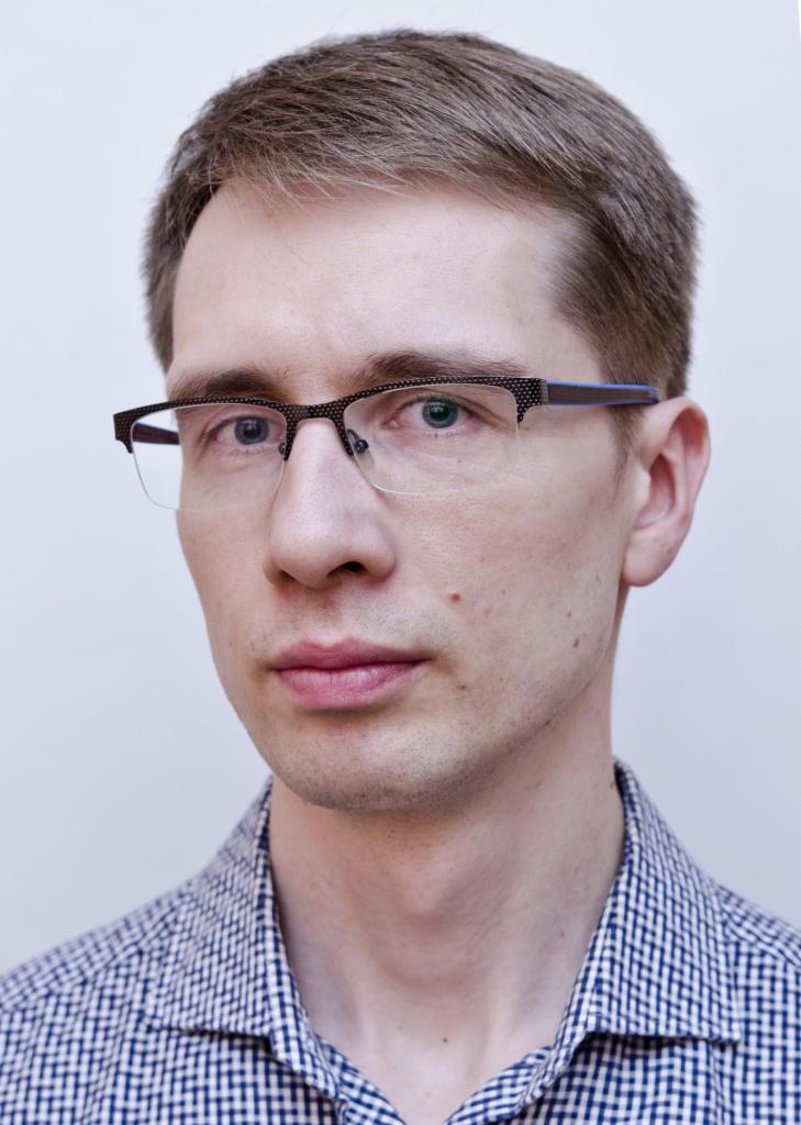 Mężczyzna w okularach z brązowymi włosami, ubrany w koszulę w kratę, na jasnym tle. Portret z bliska, skupiający się na twarzy i wyrazie.