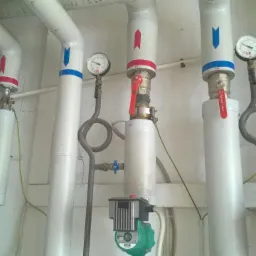 Białe rury instalacji grzewczej z zaworami, manometrami i pompami obiegowymi Grundfos, oznaczone strzałkami w kolorze czerwonym i niebieskim, na tle białej ściany.