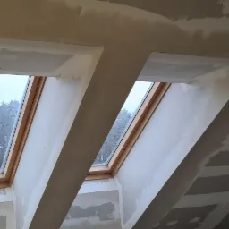 Poddasze w trakcie remontu z dwoma oknami dachowymi, widoczne płyty kartonowo-gipsowe i ślady szpachlowania na skośnym suficie