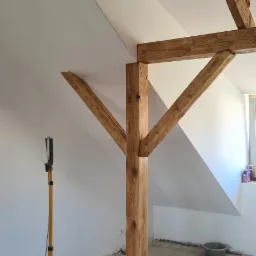 Poddasze w trakcie prac wykończeniowych z widoczną drewnianą konstrukcją nośną, lampą roboczą i workiem na śmieci.