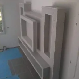 Narożna ściana z płyt karton-gips z wnękami i półkami, surowy stan wykończenia, widoczne łączenia. Podłoga częściowo zabezpieczona folią malarską.