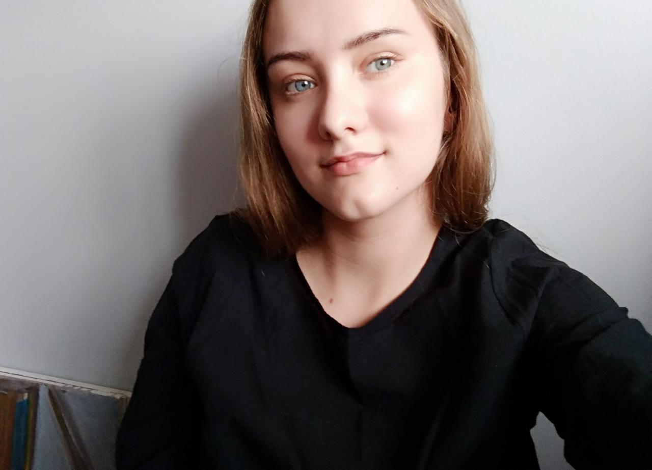 Portret młodej kobiety o jasnych włosach i niebieskich oczach, w czarnej bluzce, z delikatnym uśmiechem na tle szarej ściany. Ujęcie z góry.