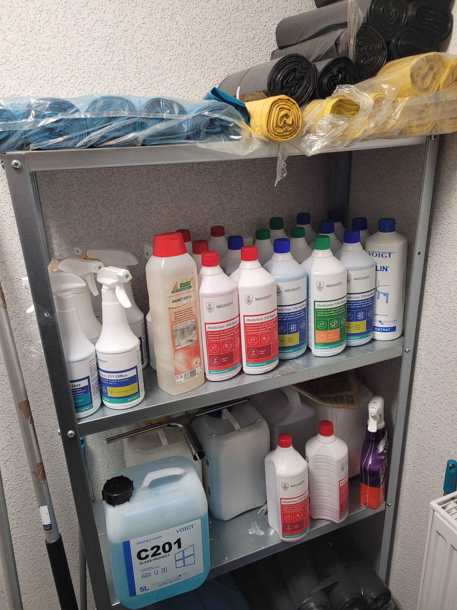 Półki regału magazynowego wypełnione środkami czystości w butelkach i kanistrach, workami na śmieci w rolkach i folią stretch, typowe wyposażenie zaplecza firmy sprzątającej.