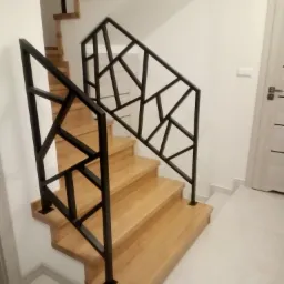 Drewniane schody z czarną, geometryczną balustradą metalową, widok z dołu, jasne wnętrze z białymi ścianami i drzwiami.