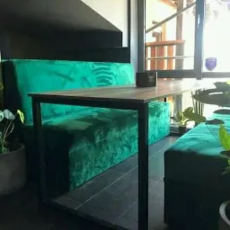 Wnętrze kawiarni z zielonymi, pluszowymi siedziskami i stolikiem z metalową ramą, widok na okno z drewnianą konstrukcją na zewnątrz.