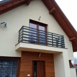 Elewacja domu z balkonem zabezpieczonym nowoczesną, poziomą, metalową konstrukcją. Widoczne drewniane elementy wykończenia, lampa zewnętrzna i fragment sąsiedniego budynku.