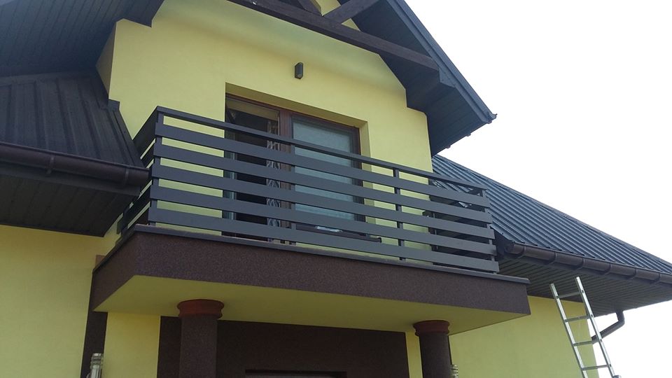 Nowoczesna balustrada balkonowa z poziomych, prostokątnych profili w kolorze grafitowym, zamontowana na balkonie domu o żółtej elewacji i ciemnobrązowym dachu, z widoczną drabiną opartą o ścianę.