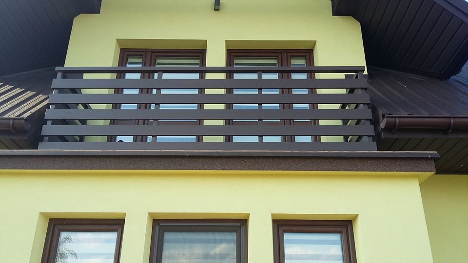 Balkon z brązową, poziomą balustradą z metalowych profili, widok z dołu na żółtą elewację budynku z brązowymi oknami i dachem.