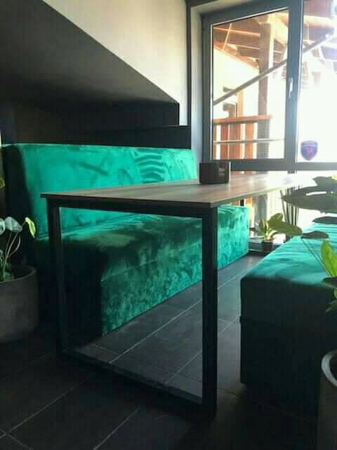 Wnętrze kawiarni z zielonymi, pluszowymi siedziskami i stolikiem z metalową ramą, widok na okno z drewnianą konstrukcją na zewnątrz.