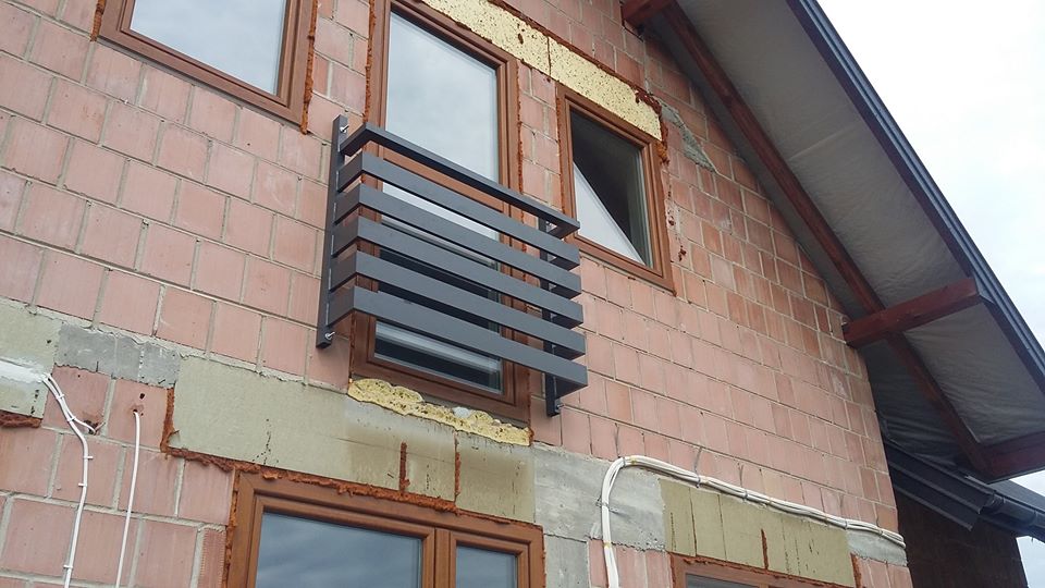 Szara, pozioma balustrada okienna zamontowana na elewacji budynku z czerwonej cegły, widoczne okna z brązowymi ramami i niezakończone prace wykończeniowe.