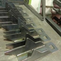 JR Modular Systems Sp. z o.o. - Rząd identycznych, metalowych elementów z przyspawanymi trójkątnymi wzmocnieniami i kwadratowymi otworami, ułożonych na metalowym stole warsztatowym, widoczne także metalowe profile.