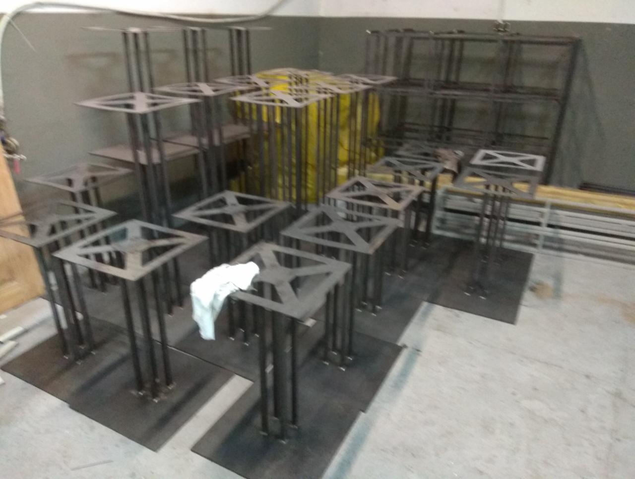 Metalowe konstrukcje spawane, przypominające podstawy stołów, ułożone w warsztacie, z widocznymi śladami obróbki i spawania, niektóre z białym materiałem do czyszczenia.