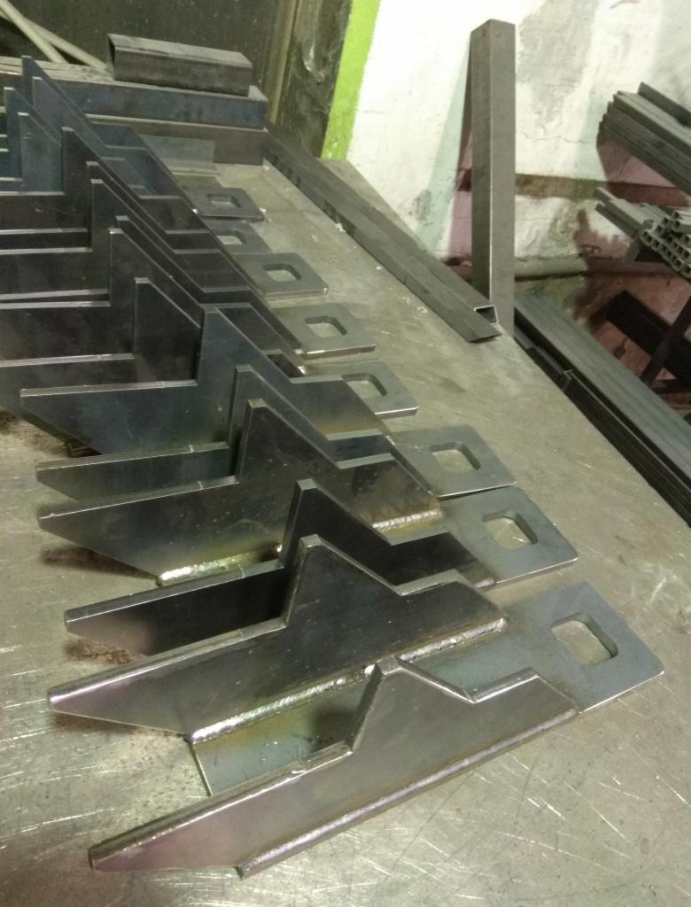 Rząd identycznych, metalowych elementów z przyspawanymi trójkątnymi wzmocnieniami i kwadratowymi otworami, ułożonych na metalowym stole warsztatowym, widoczne także metalowe profile.