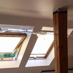 Dwa okna dachowe zabezpieczone folią malarską w trakcie prac remontowych, widoczny drewniany słup konstrukcyjny, świeżo pomalowana ściana w jasnym odcieniu.